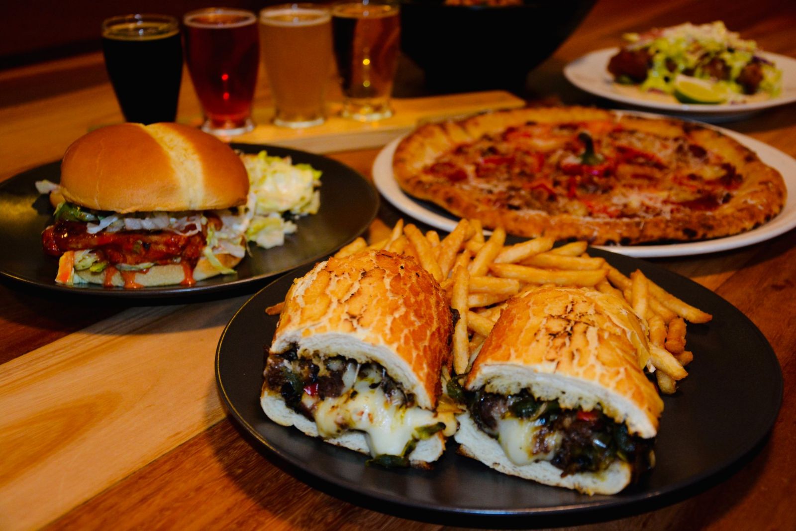 Philly cheesesteak, wood-fired pizza, a burger, a rockfish taco and a flight of house beers on a table at Rockaway Beach Brewery