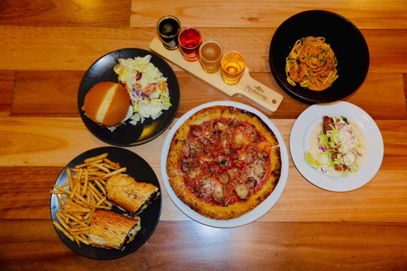 Overhead view of a Rockaway beer flight with pizza, burger, cheesesteak, pasta and a rockfish taco